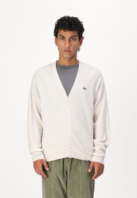 Lys beige cardigan med V-hals, fem knapper og ribbede mansjetter. Har en liten grønn logo på brystet, kombinert med en grå skjorte og grønne bukser.
