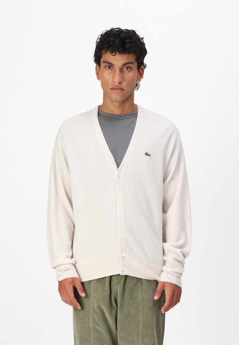 Lys beige cardigan med V-hals, fem knapper og ribbede mansjetter. Har en liten grønn logo på brystet, kombinert med en grå skjorte og grønne bukser.