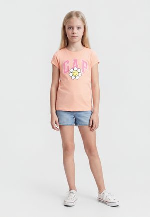 Νεαρό κορίτσι στέκεται φορώντας ροδακινί μπλουζάκι με ροζ γράμματα "GAP" και χαμογελαστή εικόνα λουλουδιού, τζιν σορτς και λευκά αθλητικά σε απλό φόντο.