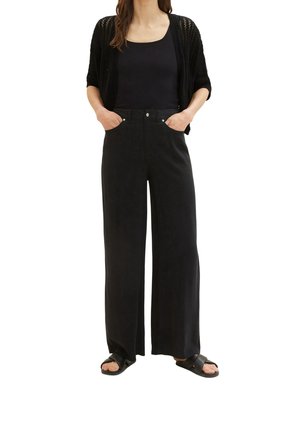 Mujer vistiendo pantalones negros de pierna ancha, top sin mangas negro, cárdigan negro de punto abierto y sandalias negras con tiras cruzadas, con las manos en los bolsillos.