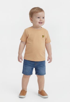 Małe dziecko stojące, ubrane w beżowy t-shirt z logo ptaka, dżinsowe krótkie spodenki i brązowe wsuwane buty, uśmiechające się i patrzące w prawo.