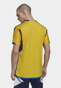 Camiseta atlética amarilla con acentos en navy, de mangas cortas y textura de malla. Presenta la etiqueta "SUERIGE" en la parte superior de la espalda y un ribete azul en el dobladillo.