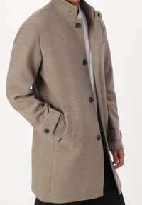 Abrigo de mezcla de lana marrón con cuello alto, cierre de botones, dos bolsillos laterales y mangas largas. Presenta un ajuste entallado y textura.
