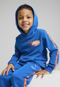 Niebieski zestaw z krojem bluzy z kapturem i spodni, wykonany z miękkiego materiału, z graficznym logo w kształcie płomienia oraz kontrastującymi czerwonymi i żółtymi paskami po bokach.