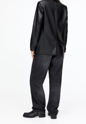 Personne portant un blazer en cuir noir, un jean gris foncé ample et des bottines noires, debout devant un fond blanc uni.