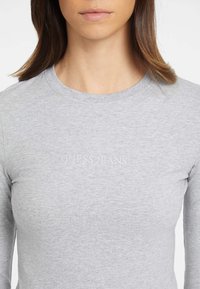 Pilkos spalvos ilgų rankovių marškiniai, pagaminti iš švelnaus medvilnės mišinio, su apvaliu iškirpimu ir tekstu „GUESS JEANS“, ryškiai išspausdintu priekyje.