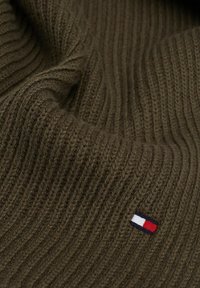 Tissu en tricot côtelé vert olive avec une texture douce. Présente un petit logo en patch rouge, blanc et bleu sur le côté.