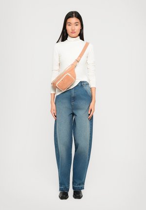 Light pink fabric crossbody bag with a zipper pocket, worn over a white turtleneck and blue high-waisted jeans. Black shoes.