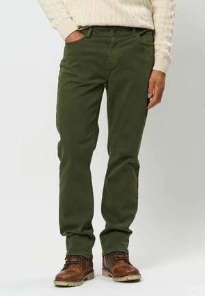 Personne portant un pantalon vert olive, des bottes en cuir marron à lacets et un pull crème en maille torsadée, debout avec une main dans la poche.