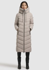 Pikk beeži puffer-jakk, mis on varustatud kapuutsi, kaldkriipsu tikandiga, lukuga ja küljetaskutega, sobitatud mustade retuusidega ja saapaga.