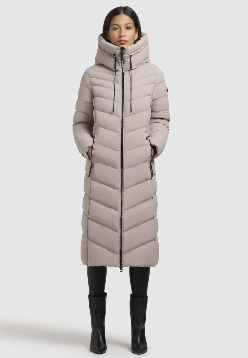 Pikk beeži puffer-jakk, mis on varustatud kapuutsi, kaldkriipsu tikandiga, lukuga ja küljetaskutega, sobitatud mustade retuusidega ja saapaga.