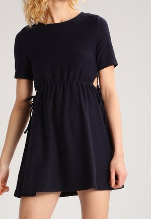 Kvinde iført en kortærmet, mørkeblå kjole med udskæringer i siderne og en taljesamling, stående foran en ensartet baggrund.