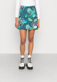 Kvinna som bär en blå blommig minikjol med en slits fram, vit långärmad topp och vita snörstövlar mot en enfärgad bakgrund.