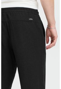 Black, lightweight pants with an elastic waist, side pockets, and a woven label on the back. Fabric shows a subtle texture.