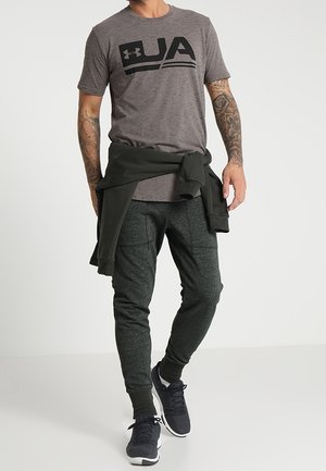 Homme portant un t-shirt gris avec le logo UA, un pantalon de jogging foncé, des baskets noires et un sweat-shirt noir noué autour de la taille, debout devant un fond blanc.