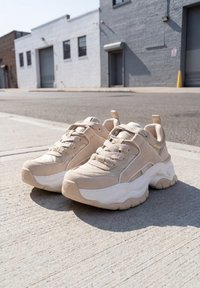 Paire de baskets beige épaisses avec des semelles blanches épaisses posées sur un trottoir en béton ensoleillé en zone urbaine.