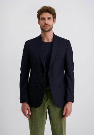 Mann mit lockigem Haar, der einen marineblauen Blazer, ein dunkles Hemd und grüne Cordhosen trägt, steht vor einem einfarbigen hellen Hintergrund.