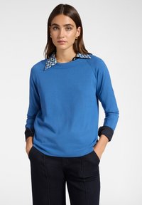 Maglione blu in morbido materiale con scollatura rotonda, dotato di un colletto a camicia patterned e polsini neri. Indossato con pantaloni scuri.
