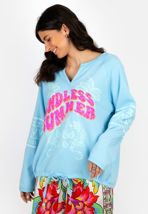 Woman wearing a light blue long-sleeve top with "Endless Summer" text and tropical designs, paired with colorful floral patterned pants.
