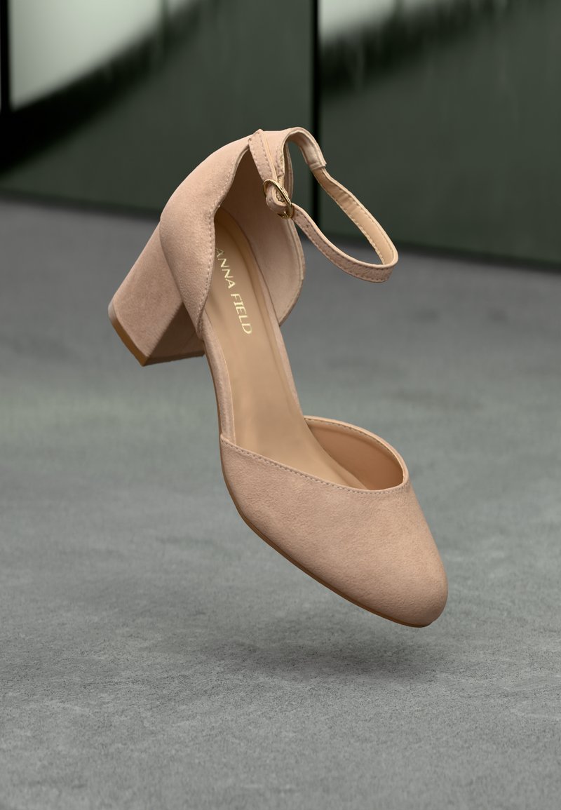 Beige nubukist tugi kõrge kontsaga king, millel on ümar varvas, pahkluu rihm ja kuldse detailiga kaunistus; sile tekstuur ja minimalism.