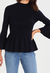 Mörkblå ribbad trikå peplum-top med hög krage, utsvängda långärmar och volangdetalj vid midjan; kombinerad med ljusblå jeans.