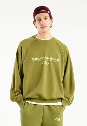 Ung mand iført en grøn New Balance sweatshirt og matchende bukser med hænderne i lommerne og en mørkerød kasket bagvendt.
