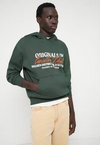 Πράσινη hoodie με μπροστινή τσέπη, λευκό και πορτοκαλί κείμενο. Φοριέται πάνω από ένα λευκό μπλουζάκι, σε συνδυασμό με ανοιχτό μπεζ παντελόνι. Απλός, καθημερινός σχεδιασμός.