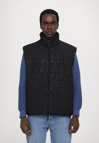 Black padded vest with a textured pattern, high collar, and zip closure, paired with a blue textured long-sleeve garment.