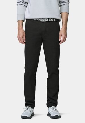 Man draagt een zwarte broek, een grijze trui, een zwart-wit patroon riem en witte sportschoenen tegen een effen achtergrond.