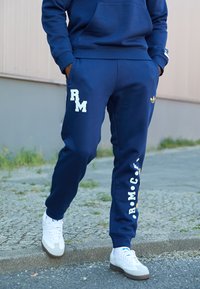 Fato de treino azul-marinho com letras brancas "RM" e "RMCF" nas calças, feito de material suave, apresenta o logótipo da Adidas numa perna. Ténis brancos.