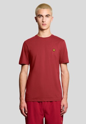 Junge Person mit kurzem blondem Haar, die ein schlichtes rotes T-Shirt und passende rote Shorts trägt, steht vor einem hellgrauen Hintergrund.