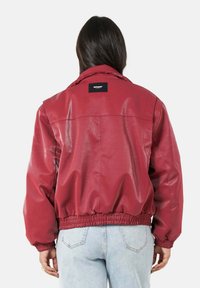 Rode nep leren jas met elastische zoom en manchetten. Heeft een omgeslagen kraag en een klein zwart label aan de achterkant. Minimalistisch ontwerp.