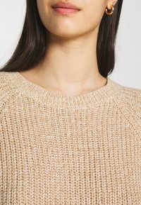 Femme portant un pull en maille beige et des boucles d'oreilles créoles dorées, montrant le cou et une partie de son visage et de ses cheveux.