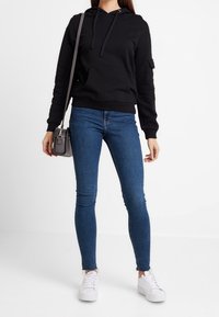 Felpa nera con cappuccio e tasca frontale, abbinata a jeans skinny blu e sneakers bianche. Una borsa con tracolla completa l'outfit.