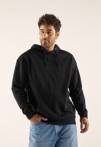 Schwarzer Kapuzenpullover aus weichem Material, mit einer Kängurutasche und Kordelzügen. Getragen mit hellblauen Jeans.