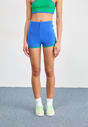 Kobieta stoi, ubrana w zielony crop top, niebieskie szorty z wysokim stanem i zielonym wykończeniem, białe skarpetki oraz jasnoniebieskie sneakersy na szarej podłodze.