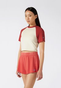 Women's two-piece outfit featuring a cream cropped top with red sleeves and red shorts with a frayed hem and logo detail.