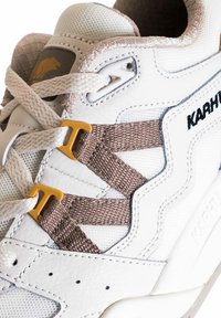 Sneaker bianco con tomaia in leather a texture e mesh, caratterizzato da cinturini in tessuto marrone e dettagli in metallo giallo. Il nome del brand "Karhu" è visibile.
