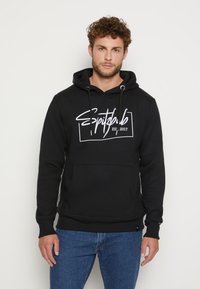 Schwarzer Hoodie mit weißem Text "Spitdub EST. 2017" auf der Vorderseite, Kordeln am Hals und einer vorderen Tasche. Getragen mit blauen Jeans.