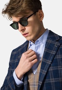 Męska ciemnoniebieska marynarka w kratę z brązowymi akcentami, jasnoniebieska koszula pod spodem oraz czarne okulary przeciwsłoneczne. Gładka faktura i dopasowany krój.