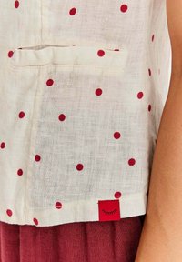 Biała tkanina lniana w czerwone kropki, z kieszonką i czerwoną metką na dolnym brzegu z motywem śpiącego oka.