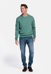 Groene gebreide trui met een ronde hals, gecombineerd met blauwe spijkerbroek en bruine suède laarzen. Model staat in een ontspannen houding tegen een witte achtergrond.