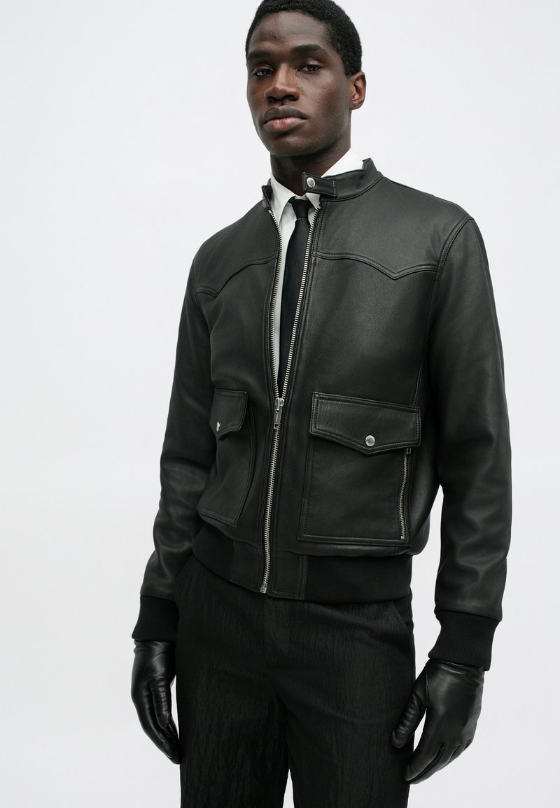 Black leather bomber jacket with a zip closure, two front pockets, ribbed cuffs, and a structured collar. Worn with black gloves and a tie.