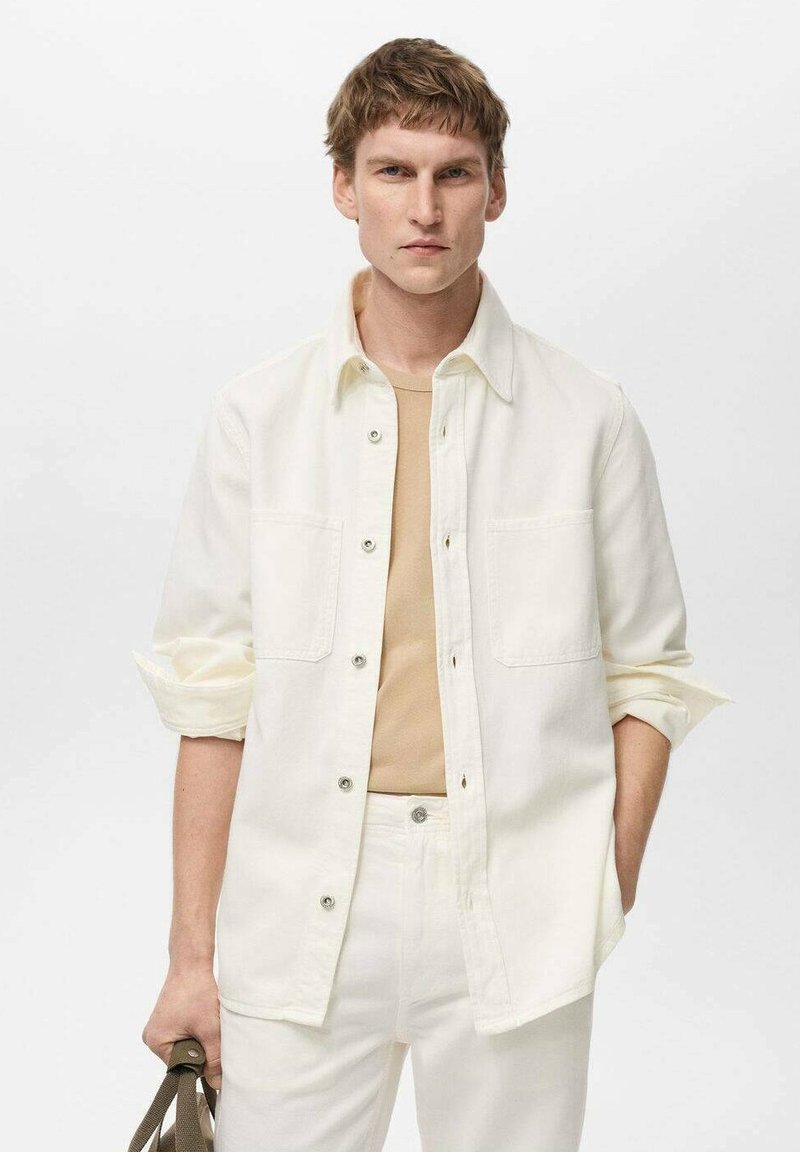 Biela košeľa na gombíky s dvoma náprsníkmi, nosená cez béžový spodný top. Model drží béžovú tašku, pričom outfit dopĺňajú biele nohavice.