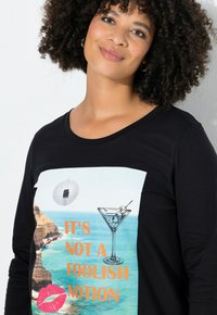 Sort langærmet skjorte med et grafisk tryk, der viser et cocktailglas, en diskokugle og tekst, sat mod en strandbaggrund.