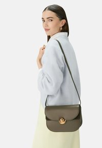 Hnědá kožená kabelka přes rameno se zlatým kruhovým zapínáním, plochý design a nastavitelným popruhem, v kombinaci se světle modrým svetr a krémovou sukní.