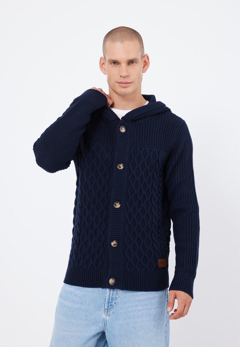 Cardigan tricotat de culoare bleumarin, cu o textură în dungi, glugă și nasturi mari. Prezintă un model în carouri și o mică etichetă din piele cu brandul.