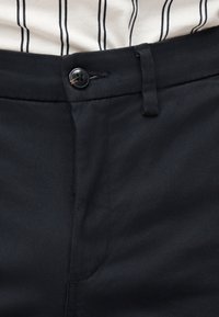 Κοντινή λήψη στη μέση μιας μαύρης παντελόνας με κούμπωμα κουμπιού και θηλιά για ζώνη, σε συνδυασμό με ένα λευκό πουκάμισο με μαύρες κάθετες ρίγες.