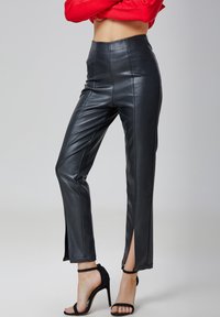 Pantalons noirs en cuir synthétique avec une taille haute, des fentes latérales à l'ourlet et une texture lisse. Associés à des sandales à talons hauts noires.