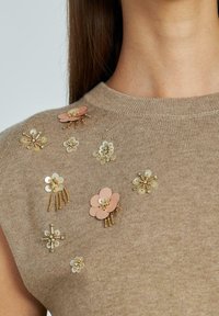 Suéter de punto beige con mangas cortas, con diversas decoraciones florales en dorado, rosa y blanco, que incluyen lentejuelas y cuentas.